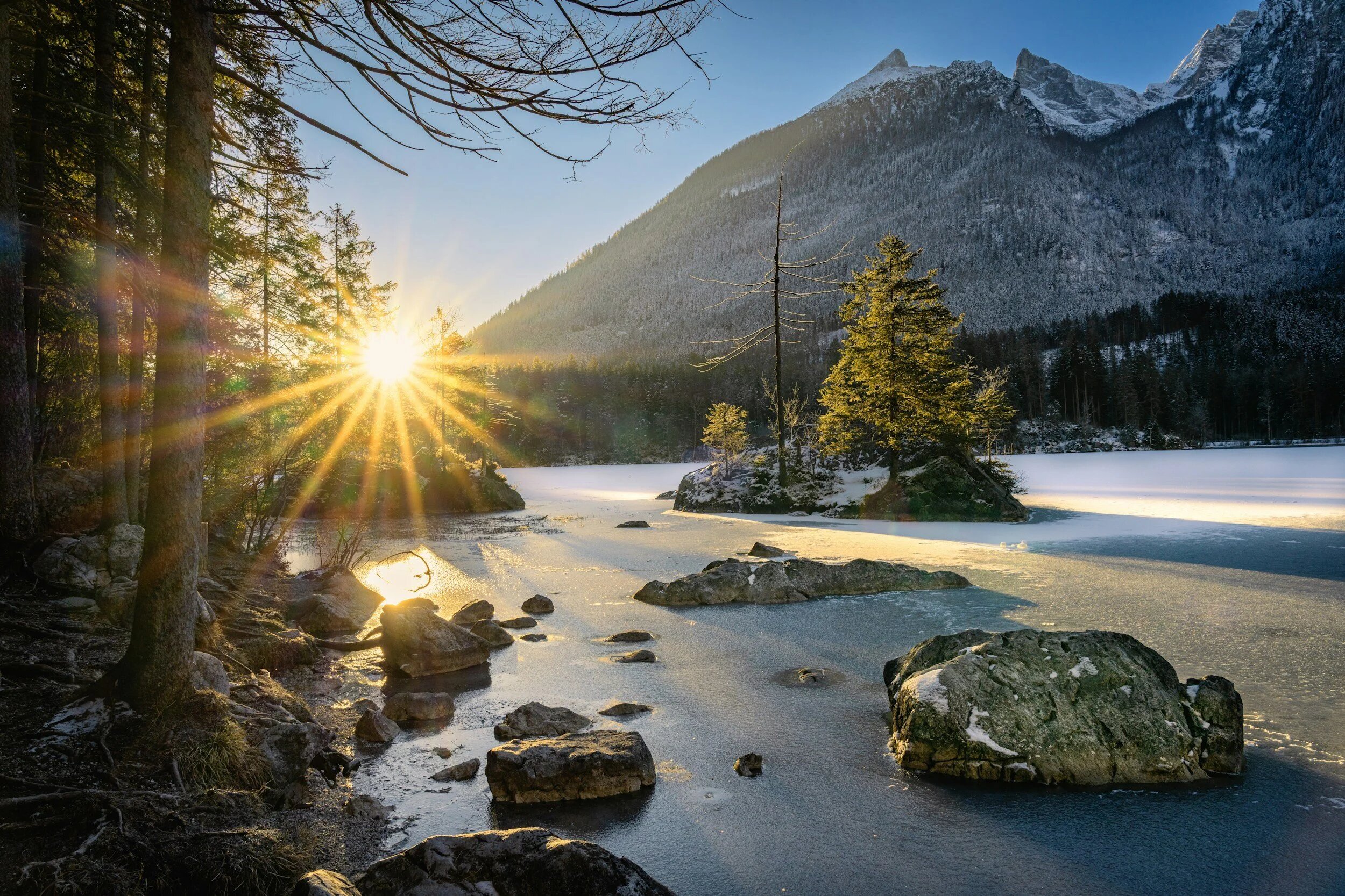 Sunrise over a still mountain lake, framed by evergreens and snow-covered peaks.